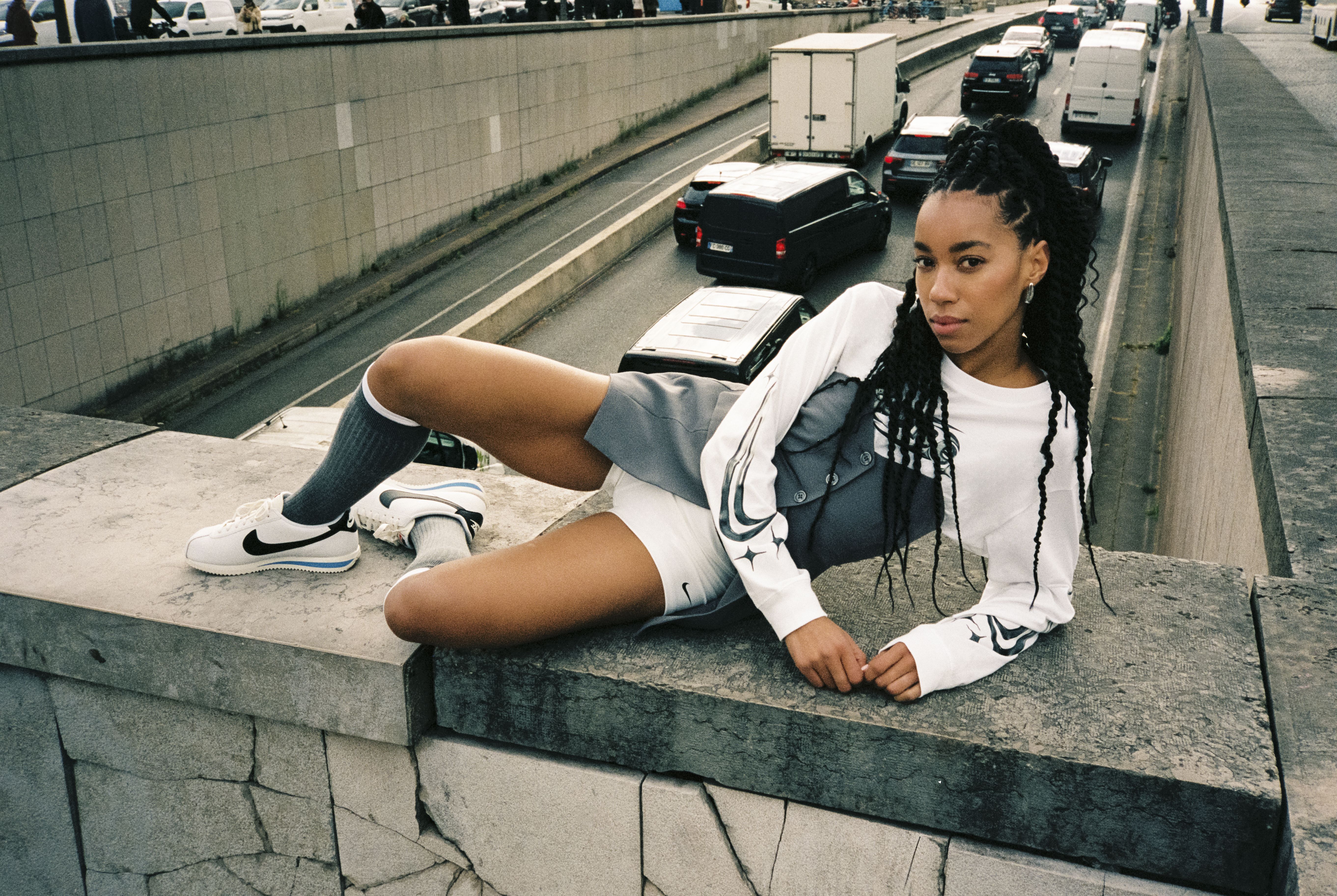 Frau im weißen NIKE-Sweatshirt und weißen NIKE-Schuhen posiert auf einer Betonmauer über einer belebten Straße.